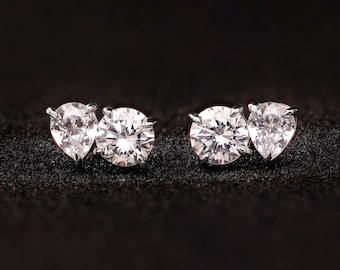 May include: A pair of stud earrings featuring a round-cut diamond paired with a pear-shaped diamond. The earrings are set in a silver metal and are displayed on a black surface. The earrings are designed to be worn as a pair.