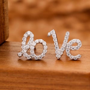 Może przedstawiać: Para srebrnych kolczyków sztyftów z napisem "love". Każda litera jest inkrustowana małymi, przezroczystymi kamieniami szlachetnymi, tworząc efekt błyszczenia. Kolczyki są prezentowane na drewnianej powierzchni.