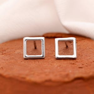 Könnte beinhalten: Ein Paar quadratische Ohrstecker aus Silber. Jeder Ohrring hat ein hohles quadratisches Design mit einem Stift zum Durchstechen. Die Ohrringe werden auf einer strukturierten, rötlich-braunen Oberfläche präsentiert.