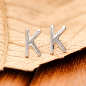 May include: A pair of silver-colored letter "K" stud earrings. The earrings are displayed on a brown leaf, set against a wooden background. The earrings are small and shiny, reflecting light.