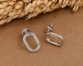 May include: A pair of silver-colored oval stud earrings. Each earring features a row of small, clear gemstones set along one side of the oval. The earrings are displayed on a brown surface, with a wheat stalk in the background.