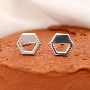 May include: A pair of silver hexagon stud earrings. Each earring has a geometric design with a solid top and an open space below. The earrings are displayed on a textured, reddish-brown surface.