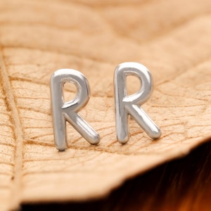May include: Two silver stud earrings, each in the shape of the letter "R". They are set against a textured, light brown background, possibly a leaf. The letters are in a rounded, sans-serif font.