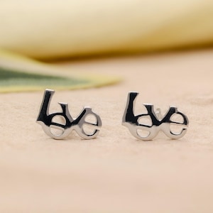 Puede incluir: Pendientes de plata con la palabra "love" escrita en cursiva. Son pequeños y delicados, con un acabado pulido. El fondo es de un color suave y neutro, resaltando los pendientes.