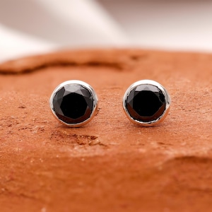 Puede incluir: Un par de pendientes de botón de piedra preciosa negra. Cada pendiente presenta una piedra negra redonda y facetada engastada en un bisel plateado. Los pendientes se muestran sobre una superficie texturizada de color marrón rojizo.