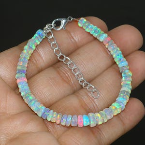 May include: A delicate bracelet featuring small, iridescent opal beads. The beads display a range of colors, including pink, blue, and green, strung together with a silver-toned chain and clasp. The bracelet is shown on a hand.