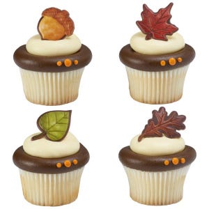 May include: Four cupcakes decorated with fall-themed toppers. The cupcakes are frosted with white frosting and have a chocolate brown bottom. The toppers include an acorn, a maple leaf, a green leaf, and an oak leaf.