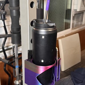Puede incluir: Un vaso térmico negro con una pajita morada se encuentra en un soporte impreso en 3D, sujeto a una máquina de pesas metálica. El soporte es morado y rosa. Hay un bolígrafo en el soporte. El vaso es para bebidas frías o calientes.