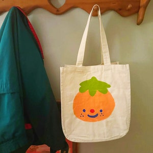May include: A white canvas tote bag with a hand-painted orange fruit design. The fruit has a green stem and a smiling face with blue eyes and a red nose.