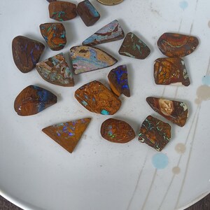 Könnte beinhalten: Eine Sammlung von 15 kleinen, unregelmäßig geformten Opal-Steinen in Braun-, Blau- und Grüntönen. Die Steine sind auf einem weißen Teller mit einem dekorativen Muster angeordnet.