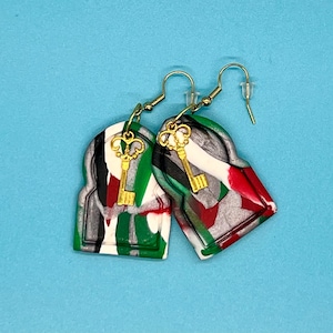 Puede incluir: Un par de pendientes verdes, blancos, rojos y plateados con un dije de llave dorada. Los pendientes tienen forma de ojo de cerradura.