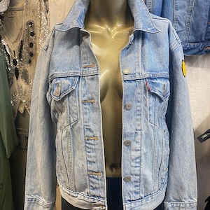 May include: Light blue denim jacket with a classic collar, button-up front, and two chest pockets. Features a small red tag and a yellow patch on the sleeve. The jacket is displayed on a mannequin.