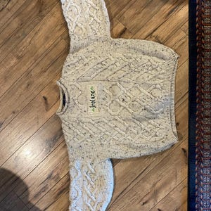 May include: A cream-colored cable knit sweater featuring a speckled texture throughout the fabric. The garment has long sleeves, a crew neckline, and a rectangular patch on the chest with the word Ireland and a small green shamrock icon.