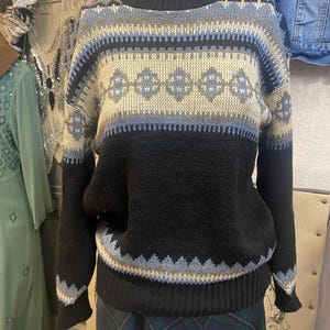 May include: A black sweater with a geometric pattern in cream, light blue, and gray. The sweater has a crew neck and long sleeves. The pattern includes diamond shapes and horizontal stripes. The bottom and cuffs are ribbed.