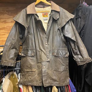 May include: Brown waxed cotton jacket with a corduroy collar and shoulder cape. The jacket has multiple pockets, a button-up front, and a quilted lining. The brand name "Kakadu" is visible on a tag inside the jacket. The jacket is hanging on a wooden hanger.