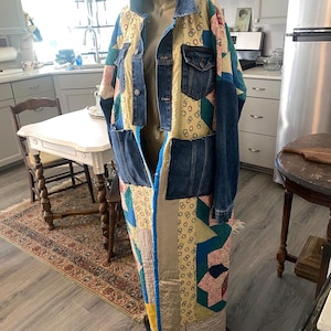 May include: A long coat constructed from a combination of blue denim fabric and multi-colored patchwork quilt panels. The garment features a denim collar, button-front closure, two chest pockets, and long sleeves, with a hem reaching below the knees.