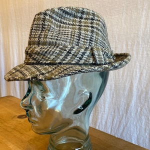 May include: A brown and black plaid fedora hat with a wide brim. The hat is on a glass mannequin head.