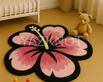 May include: A pink and black hibiscus flower-shaped rug with a yellow stamen. The rug is on a light wood floor, near a crib and a teddy bear. The text "YAZKNOT" is visible on the rug.