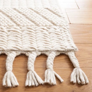 May include: A white woven rug with a geometric pattern and fringe. The rug is made of natural fibers and has a soft, textured feel.
