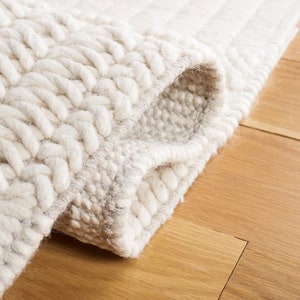 May include: Close-up of a white and cream-colored woven rug with a textured, braided design. The rug is folded over, showing the underside of the weave.