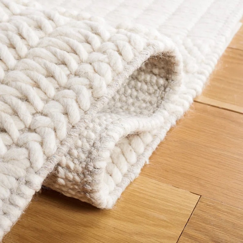 May include: Close-up of a white and cream-colored woven rug with a textured, braided design. The rug is folded over, showing the underside of the weave.