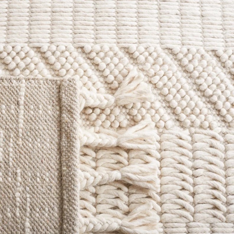May include: A white woven rug with a textured pattern and fringe. The rug has a subtle beige border.