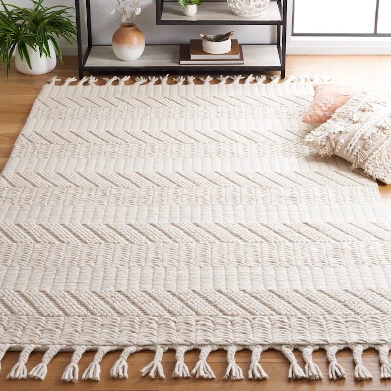 May include: A cream-colored woven rug with a geometric chevron pattern and fringe edging. The rug is made of natural fibers and has a soft, textured feel.