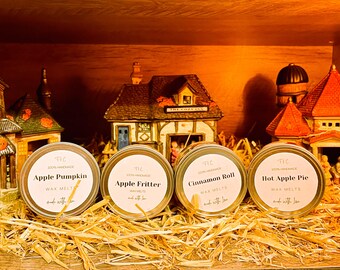 May include: Four round wax melt tins with labels reading "Apple Pumpkin," "Apple Fritter," "Cinnamon Roll," and "Hot Apple Pie." The tins are on a bed of straw, with miniature village houses in the background. The text "100% Handmade" is visible on the labels.