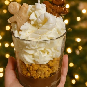 Op de afbeelding: Een kaars in een glas, die op een dessert lijkt, wordt in een hand gehouden. De kaars heeft lagen bruine, oranje en witte was, bekroond met peperkoekmannetjes. De achtergrond is vervaagd met bokeh-lichten.
