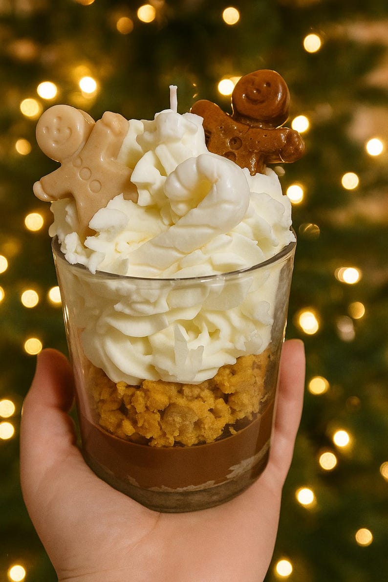 Op de afbeelding: Een kaars in een glas, die op een dessert lijkt, wordt in een hand gehouden. De kaars heeft lagen bruine, oranje en witte was, bekroond met peperkoekmannetjes. De achtergrond is vervaagd met bokeh-lichten.