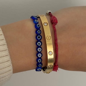 Może przedstawiać: Zestaw trzech bransoletek: niebieska bransoletka z koralików z motywem oka proroka, złota metalowa bransoletka typu bangle ze śrubkami i osadzonymi przezroczystymi kamieniami oraz cienka czerwona bransoletka ze sznurka z małym białym koralikiem.