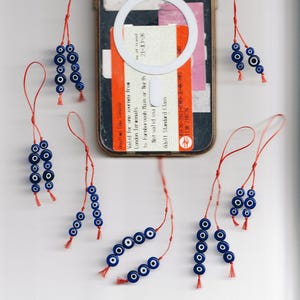 May include: A collection of blue and white evil eye charms on red strings, arranged around a smartphone with a white circular magnetic ring and a train ticket insert. The charms feature circular glass beads with concentric blue and white rings.