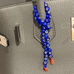 May include: A blue beaded charm with white and black eye designs hangs from a gray leather handbag. The charm has a tassel of orange threads at the bottom. The handbag has silver hardware.