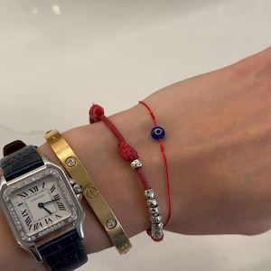 Puede incluir: Un reloj plateado con correa de cuero azul oscuro, un brazalete dorado con piedras transparentes incrustadas, una pulsera de cordón rojo con cuentas plateadas y una pulsera de hilo rojo fina con un pequeño dije de ojo turco azul y blanco.