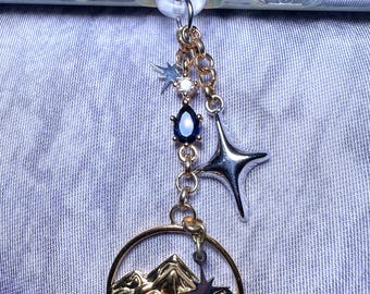 May include: A gold charm with a mountain scene and a star. The charm is attached to a gold chain with a blue gemstone and a silver star.