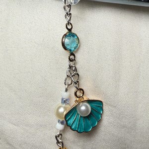 Peut inclure: Un charme en forme de coquille de couleur turquoise avec une perle à l'intérieur, suspendu à une chaîne argentée avec des perles blanches et bleues, une gemme turquoise et une perle dorée.