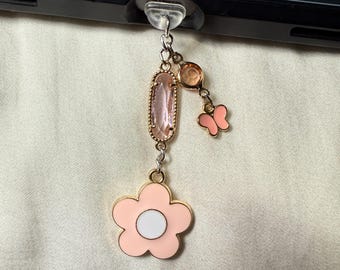 May include: A pink flower charm with a white center, a pink butterfly charm, and a pink and gold crystal charm hanging from a gold chain. The charms are attached to a clear plastic phone charm holder.