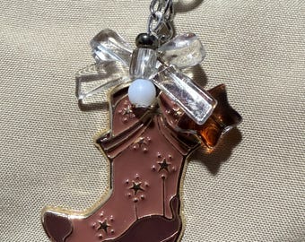 May include: A pink cowboy boot charm with gold trim and stars. The charm is attached to a silver chain with a clear glass bow and a brown star.
