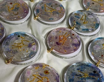 May include: A collection of round, clear resin pendants with gold constellation designs. Each pendant features a unique blend of colors, including purple, blue, and gold flakes. They have a gold-colored charm and a white base.