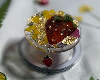 May include: A clear, round resin badge reel with a strawberry and yellow flower design, accented with gold leaf. A small strawberry charm hangs from the reel. The reel is sitting on a white surface.