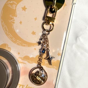 May include: A gold-colored keychain with a heart-shaped clasp and a charm featuring a mountain landscape and stars. The charm is attached to a chain with blue and silver star accents. The background is a phone case with a moon and stars design.