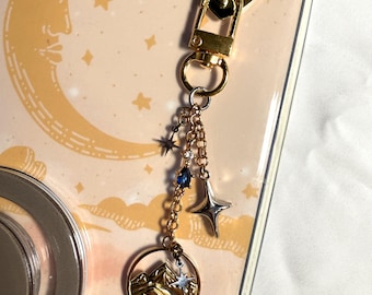May include: A gold-colored keychain with a heart-shaped clasp and a charm featuring a mountain landscape and stars. The charm is attached to a chain with blue and silver star accents. The background is a phone case with a moon and stars design.