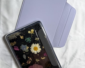 May include: A clear phone case with a black background, embedded with dried flowers in shades of pink, white, yellow, and blue, and gold leaf accents. A light purple tablet case is behind the phone case.