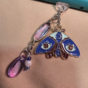Peut inclure: Un charme représentant un papillon de nuit bleu et brun avec une lune croissante et des étoiles sur ses ailes. Le charme est attaché à une chaîne argentée avec une perle de verre violette et transparente et une perle blanche.