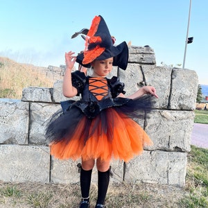 May include: A young girl dressed as a witch for Halloween. She is wearing a black and orange tulle dress with a black sequined bodice and a black witch hat with orange accents. She is also wearing black tights and black shoes.