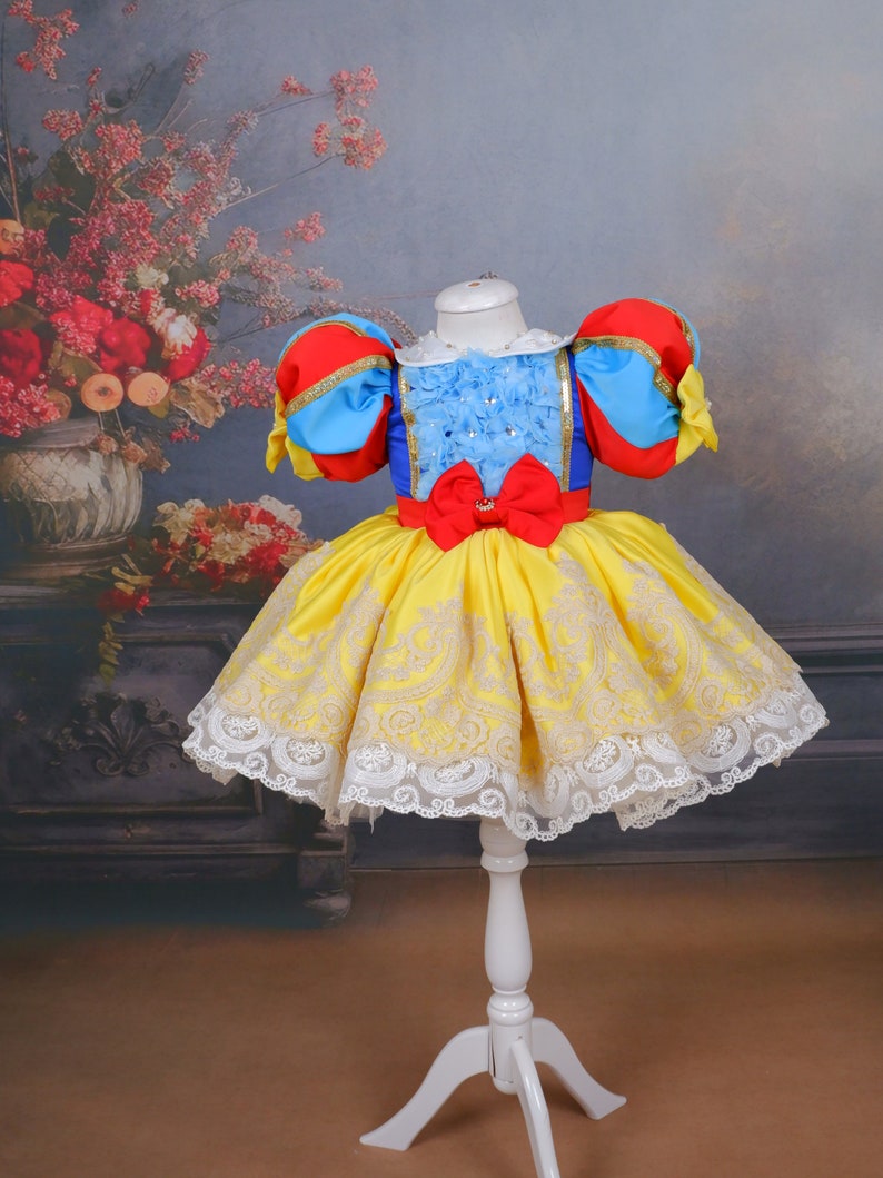 Op de afbeelding: Een gele jurk met witte kant en rode, blauwe en witte pofmouwen. De jurk heeft een rode strik in de taille en is versierd met gouden borduurwerk.