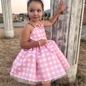 May include: A young girl wearing a pink and white gingham dress with a tulle underskirt. She is wearing a pink crown headband and a colorful beaded necklace. The girl is standing next to a white pillar.