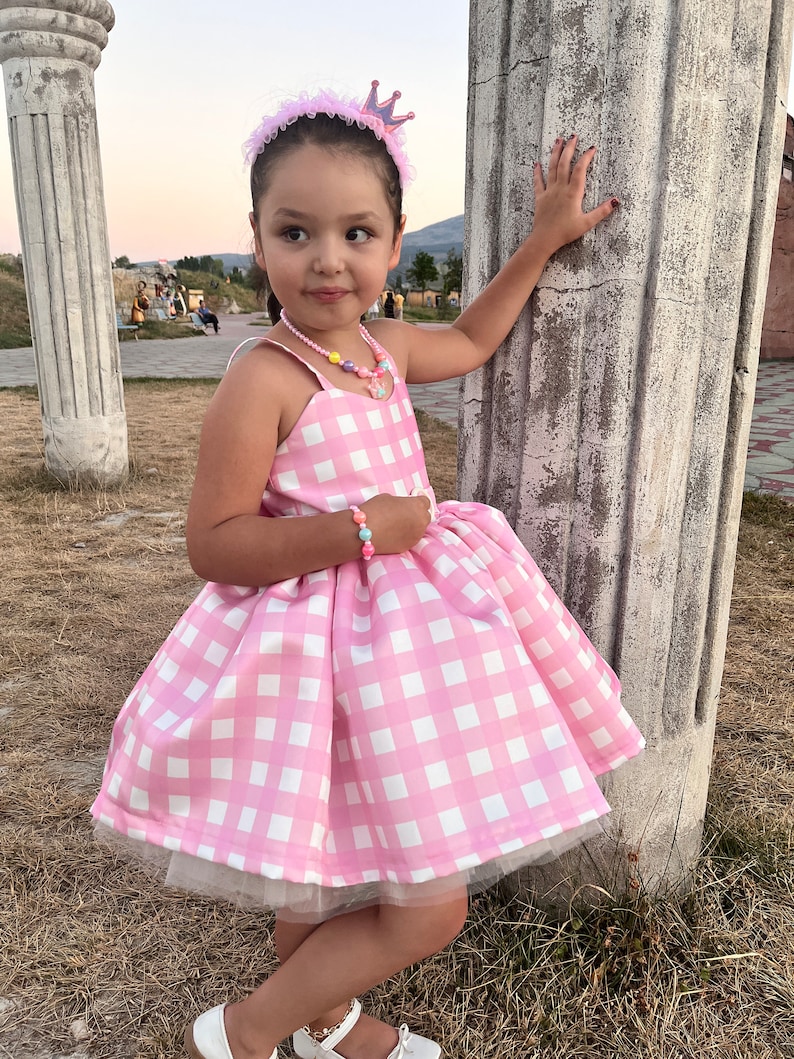 May include: A young girl wearing a pink and white gingham dress with a tulle underskirt. She is wearing a pink crown headband and a colorful beaded necklace. The girl is standing next to a white pillar.