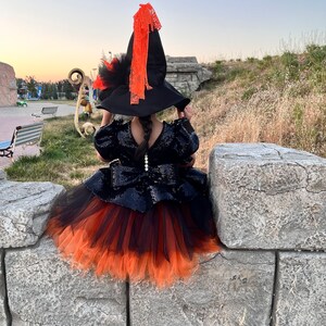 May include: A young girl dressed as a witch for Halloween. She is wearing a black and orange tulle skirt, a black sequined top, and a black witch hat with an orange ribbon.