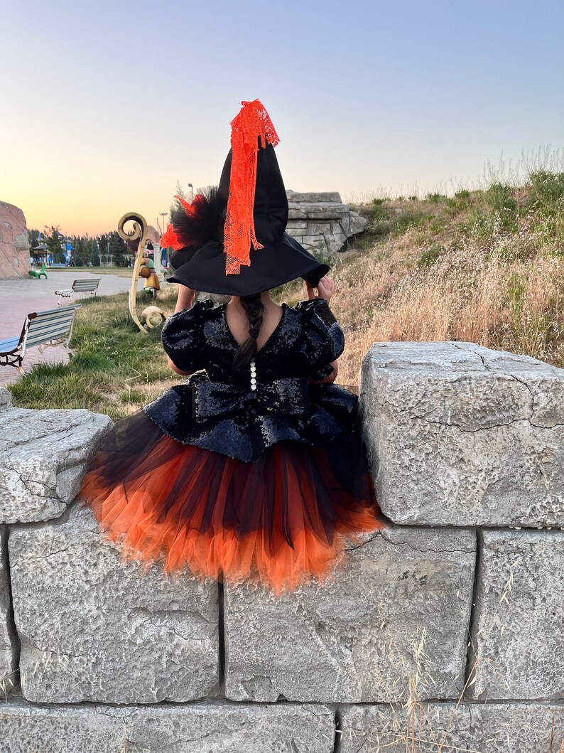 May include: A young girl dressed as a witch for Halloween. She is wearing a black and orange tulle skirt, a black sequined top, and a black witch hat with an orange ribbon.